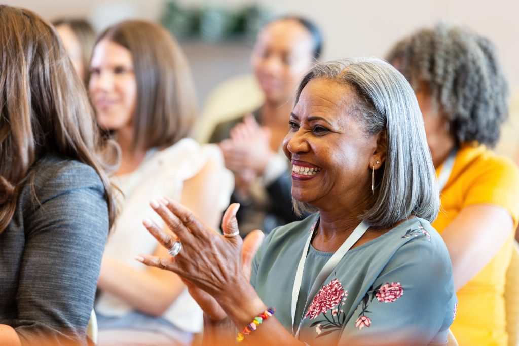 Close-up of smiling women clapping enthusiastically. They are seated indoors at an event, suggesting shared engagement and enjoyment.