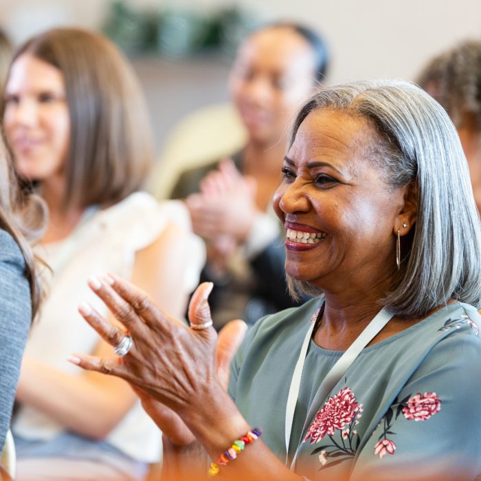 Close-up of smiling women clapping enthusiastically. They are seated indoors at an event, suggesting shared engagement and enjoyment.