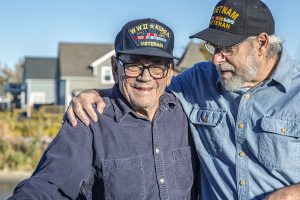 Two generations of real person senior adult men USA military veterans. On the left, a 93 year old United States Army World War II and Korean Conflict US Air Force military veteran is smiling at the camera while his son-in-law on the right - a 65 year old US Navy sailor Vietnam War military veteran - is teasing him - trying to distract him by telling jokes during the photo shoot. They are both wearing generic souvenir shop military veteran commemorative baseball style caps.